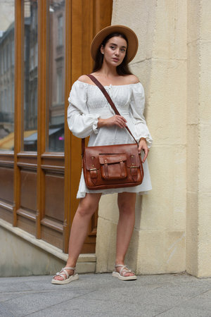 brunette with long hair wearing white romantic summer dress and beige hat posing with luxury leather bagの写真素材