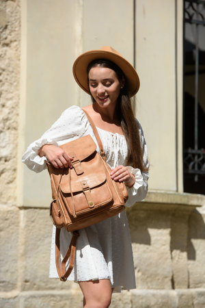brunette with long hair wearing white romantic summer dress and beige hat posing with luxury leather backpackの写真素材