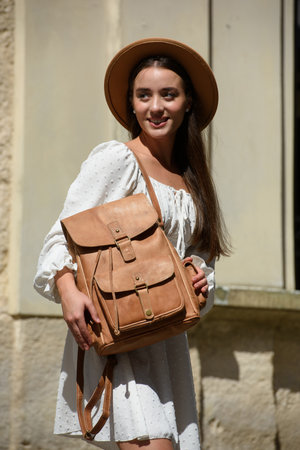 brunette with long hair wearing white romantic summer dress and beige hat posing with luxury leather backpackの写真素材