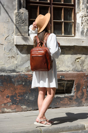 brunette with long hair wearing white romantic summer dress and beige hat posing with luxury leather backpackの写真素材