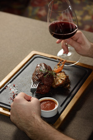 man sitting at a table in a restaurant. He is holding a glass of red wine and eating a steakの写真素材