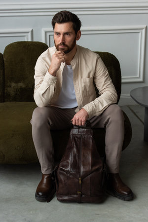 A man is comfortably sitting on a sofa, with a leather brown backpackの写真素材