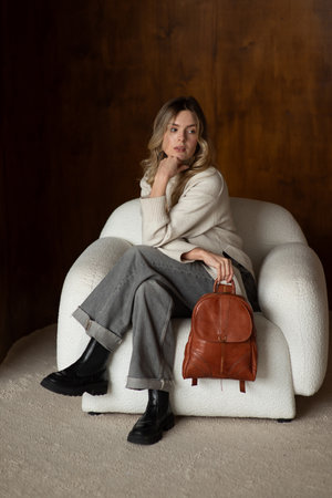 beautiful girl in gray pants and beige sweater posing with a small shiny brown leather backpackの写真素材