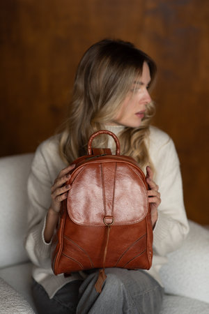 beautiful girl in gray pants and beige sweater posing with a small shiny brown leather backpackの写真素材