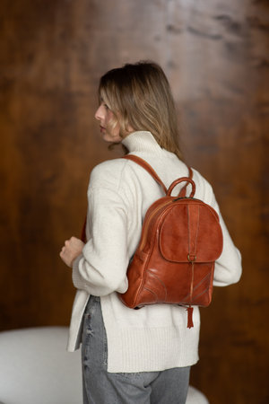 beautiful girl in gray pants and beige sweater posing with a small shiny brown leather backpackの写真素材