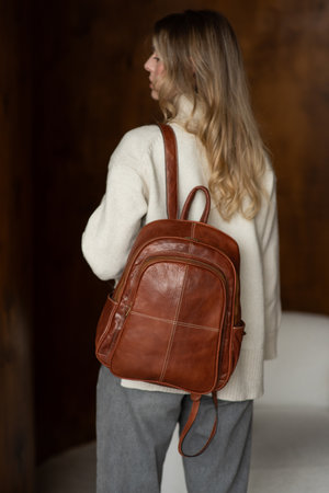 beautiful girl in gray pants and beige sweater posing with a small shiny brown leather backpackの写真素材