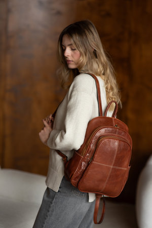beautiful girl in gray pants and beige sweater posing with a small shiny brown leather backpackの写真素材