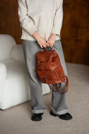 beautiful girl in gray pants and beige sweater posing with a small shiny brown leather backpackの写真素材