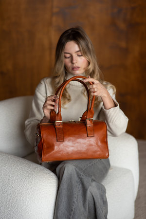 beautiful girl in gray pants and beige sweater posing with a small shiny brown leather bagの写真素材