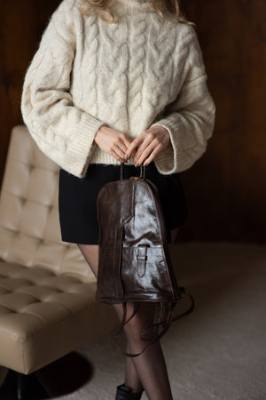 A girl in a warm sweater, a black short skirt and nylon tights is posing with a brown backpack.の写真素材