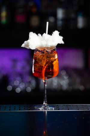 A vibrant orange cocktail filled with ice sits on a textured bar, topped with a whimsical cloud of white cotton candy and a white straw.の写真素材