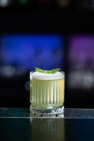A pale yellow cocktail with a frothy white top is garnished with two vibrant green mint leaves, sitting on a reflective bar surface.の写真素材