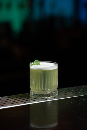 A pale yellow cocktail with a frothy white top is garnished with two vibrant green mint leaves, sitting on a reflective bar surface.の写真素材
