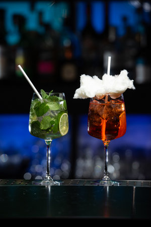 Two uniquely garnished cocktails on a reflective bar surface, one brimming with fresh mint and a straw, the other a vibrant orange hue topped with fluffy white sugar cotton candy and a white strawの写真素材