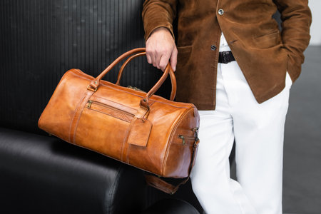 Close up of a man holding a premium tan leather duffel bag wearing a suede blazer and white trousers