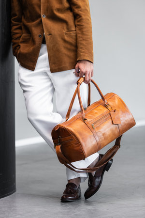 Close-up of a man holding a premium brown leather duffel bag wearing a suede blazer and white...