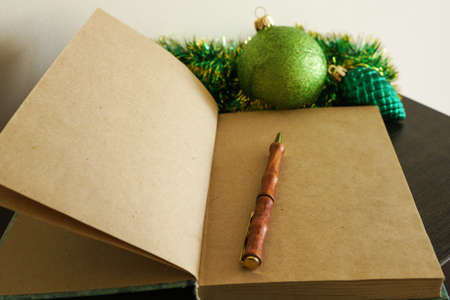 An open blank notebook with aged brown sheets and a brown pen in the form of a tree trunk, against a background of green Christmas tinsel and a green Christmas tree shiny ball in sequinsの写真素材