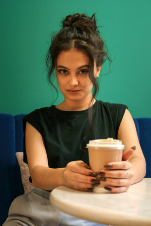 A girl sits at a table in a cafe with a glass of coffee, looks at the camera and smilesの写真素材