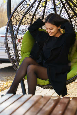 A beautiful girl sits on a terrace near a cafe in a hanging chair, she smiles and straightens her...