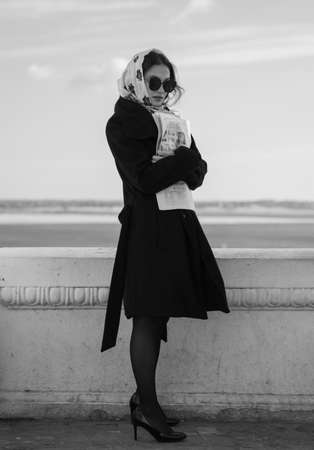 A model stands on the embankment and hugs a newspaper, wearing black glasses and a black coatの写真素材