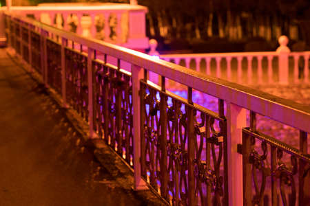 Night photograph from the bridge to the pond, the ornate cast iron railing of the bridge in the foreground under the pink colored lighting of the park lightsの写真素材