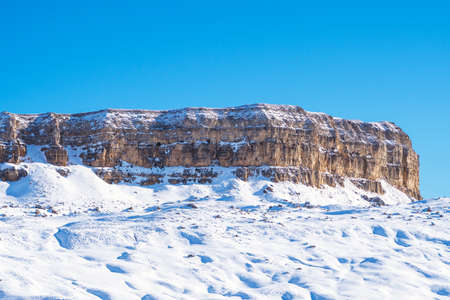 View of a small snow-covered chalk mountain, in rare places overgrown with trees against a blue skyの写真素材