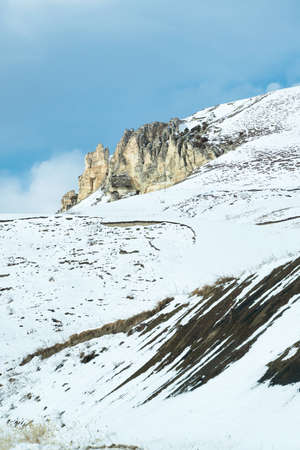 View of a small snow-covered chalk mountain, in rare places overgrown with trees against a blue skyの写真素材