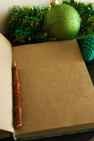 An open blank notebook with aged brown sheets and a brown pen in the form of a tree trunk, against a background of green Christmas tinsel and a green Christmas tree shiny ball in sequinsの写真素材