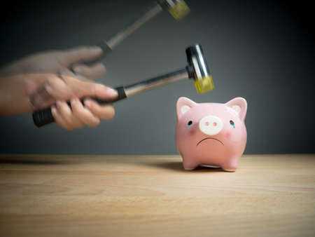 A hand holding a hammer which is raised above a pink sad piggy bank, with a shocked and apprehensive facial expression. Reflective surface and light grey background.の写真素材