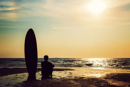 Silhouette of surf man sitting with a surfboard on the seashore beach at sunset time. Handsome Asia man model in his 20s.の写真素材