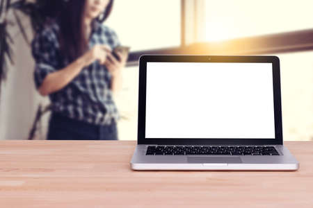Office workplace with blank white screen laptop with clipping path on wood table. Woman use mobile phone by touch at background near window. Asian women modelの写真素材