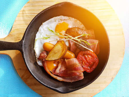 Breakfast in cooking pan with fried eggs, sausages, bacon , tomato , potato and vegetable on wood block background on the table in the kitchen. morning light effectの写真素材