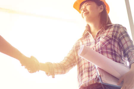 happy Asian woman engineer as construction workers with blueprint and contract in hand with protective yellow helmet are shaking hands while working at construction siteの写真素材