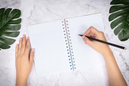 flat lay of woman hand writing in a blank white page notebook on white marble desk, top view. green...