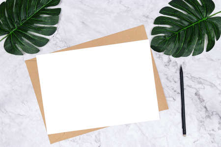 blank white paper size A4 on brown paper envelope on white marble background with green palm leaf on...