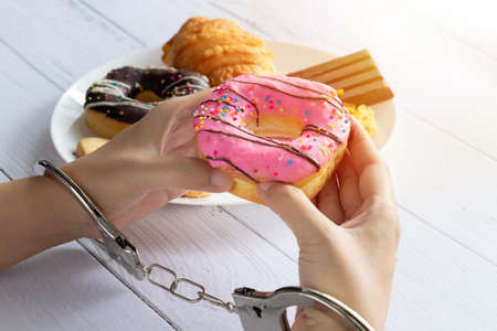 Calories control, food diet and weight loss concept. top view of two hands was control by handcuff, holding doughnut from white dish on dining tableの写真素材