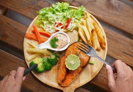 salmon steak served on wooden plate ,side dishes are boiled vegetables , French fries , fresh salad ,cream sauce and piece of lemon on topの写真素材