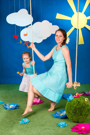 Mother with small daughter on the swing on the blue background with sun and cloudsの写真素材