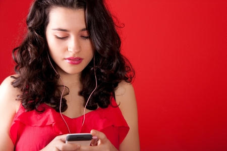 Cute young woman choosing a song or a radio station in her mobile phone. Wearing pink earphones. Red background.の写真素材