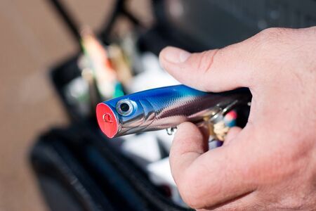 Blue fishing lure in fisherman hand and sand and a bag with more lures out of focus at the background の写真素材