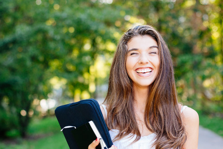 Portrait of a laughing student girl with eyes closed holding a laptop outdoors