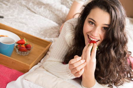 Beautiful young woman having breakfast in bed and eating strawberriesの写真素材