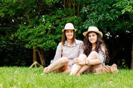 Cute teen girl friends with hats sitting on the grass having good momentsの写真素材