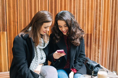 Two beautiful and happy young women using a mobile phone while having a coffee in a cafeの写真素材