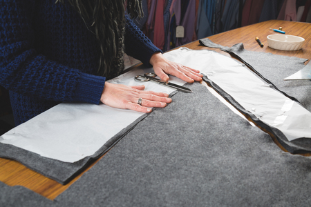 Hands of seamstress cutting a grey fabric with scissors using a paper template
