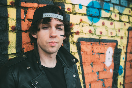 Portrait of a young man with leather jacket and a cap with a black painted stripe in his cheek looking at camera in font of a graffiti wallの写真素材