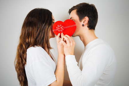 Beautiful young couple about to kiss behind a red heart shaped gift box on white backgroundの写真素材