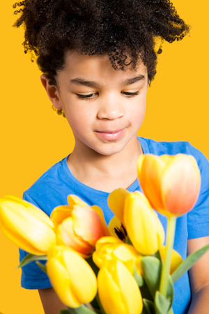 Adorable little boy with a blue t-shirt holding a tulips bouquet. Isolated on yellowの写真素材