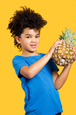 Happy little boy with a blue t-shirt holding a pineapple. Isolated on yellowの写真素材