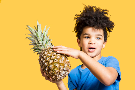 Happy little boy with a blue t-shirt holding a pineapple. Isolated on yellowの写真素材